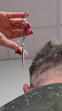 Barber cutting a gray-haired man's hair with scissors and comb from a low-angle view. Vertical close-up video barber trimming mature man hair inside barbershop or beauty salon.