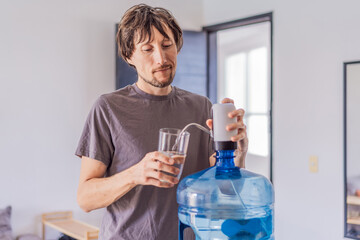 Man filling a glass with drinking water from a large 20-liter water dispenser with an electric...