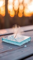 A delicate white feather rests on a square glass block, set on a wooden surface, with a blurred, golden background suggesting sunlight.