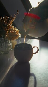 Woman&rsquo;s hands pour tea from a ceramic teapot into a cup on a kitchen table, backlit by soft autumn sunrise, creating a peaceful, meditative morning atmosphere.
