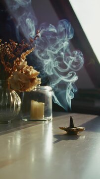 9x16 vertical close-up of smoke rising from a burning incense cone by a sunlit window on an autumn morning, with flowers on the table creating a serene, meditative, calming mood.