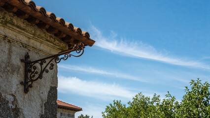 A building corner with a decorative wrought iron bracket, roof with tiles, blue sky with wispy clouds, and green foliage.