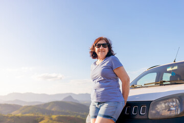 Brazilian mature woman enjoying a peaceful road trip, smiling beside her car with a scenic mountain view. Inspiring independence, freedom, and confidence for women over 60