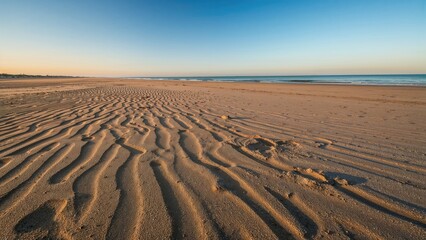 Naklejka premium A sandy beach with rippled patterns, clear blue sky, and calm ocean waves. Nature and landscape, tranquility and serenity, outdoor scene.