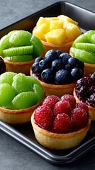 Close-up of a variety of fruit tarts on a dark tray. The tarts are filled with different fruits, including kiwi, blueberries, raspberries, and mango. The lighti