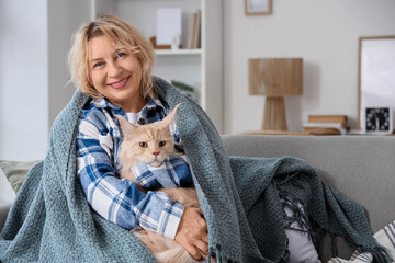 Beautiful middle-aged woman with cute cat and warm blanket sitting on sofa at home