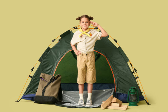Little girl scout with tent and camping equipment making scout salute on green background. World Scout Day
