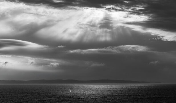 Lonely sailboat under dramatic moody storm clouds and sun rays over black and white seascape minimalist ocean horizon spiritual light over water fine art seascape photography of hope and solitude