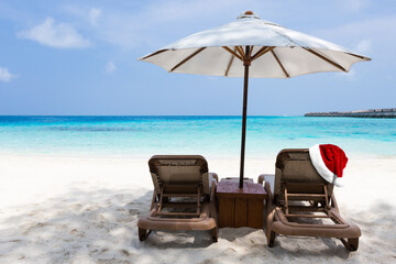 Santa Claus hat on tan chairs under white parasol on turquoise sea beach, perfect vacation on Maldives island. Christmas and New Year celebration