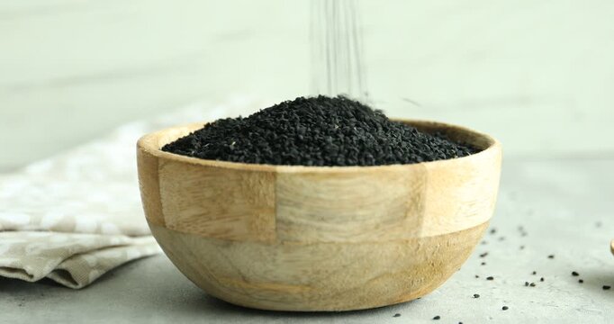 Pouring aromatic black caraway into wooden bowl at grey table, closeup