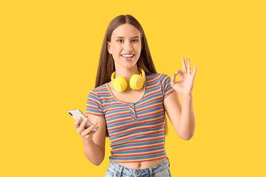 Beautiful young woman with modern mobile phone and headphones showing OK on yellow background