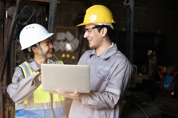 Smiling factory engineers review plan on laptop in industrial workshop with safety helmet and reflective vest under warm light, teamwork and confidence show progress and care