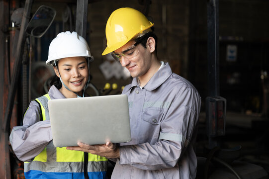 Smiling engineers review laptop in factory, industrial teamwork planning under hard hat safety gear near forklift, confident collaboration and quality control on site