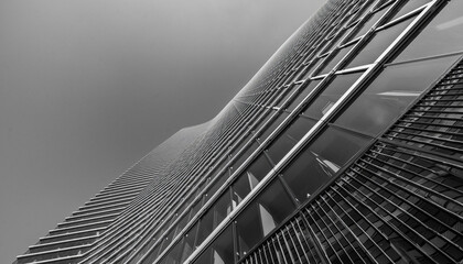 Low-angle black and white shot of a modern skyscraper's exterior with a geometric pattern of windows and metal beams against a cloudy sky.