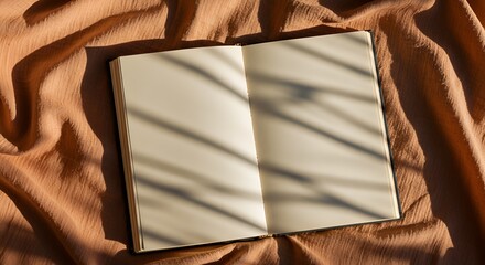 An open blank book rests on textured, earthy toned fabric illuminated by dynamic light and shadow.