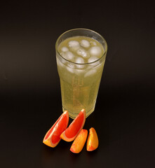 Tall faceted glass of fruit juice with ice on a black background, next to it are pieces of ripe peach and grapefruit.