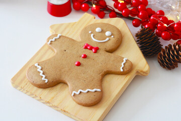 A single, smiling gingerbread man cookie with white icing and a red bow tie, presented on a wooden cutting board with Christmas decorations nearby.
