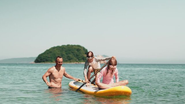 Family enjoys sunny day paddleboarding in calm turquoise water. Parent guides two kids as they balance on board, creating joyful moment filled with summer energy and playful outdoor fun.
