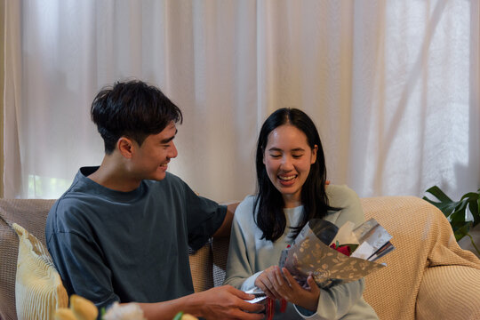 Happy young couple love home, man gives woman romantic valentine day flowers and gift, celebrating special occasion, enjoying joyful connection with warm affection