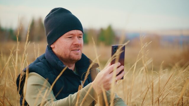 A man dressed in warm clothing sits in a field of tall grass while using a smartphone, enjoying a moment of connection with nature and technology in a tranquil outdoor setting.