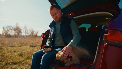A Man Enjoys a Relaxing Moment with His Dog by the Car in a Scenic Outdoor Setting, Embracing Nature While Sipping a Warm Beverage Under the Beautiful Sky