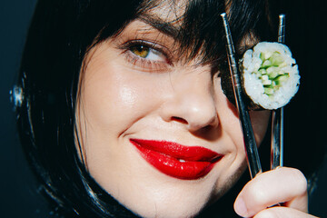 Young woman with red lips holding sushi with chopsticks, striking a playful pose against a dark background, focusing on food and enjoyment.