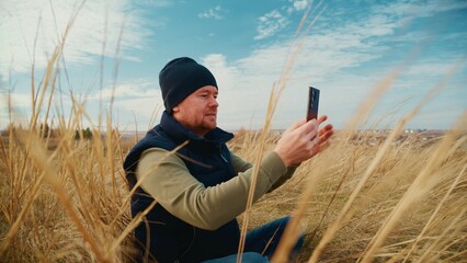 A Man Sitting in a Field of Tall Grass, Engaged in the Activity of Using a Smartphone, Surrounded by an Expansive Sky and Nature in a Tranquil Setting