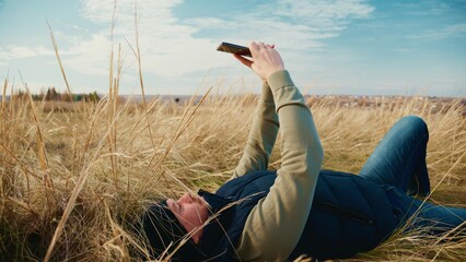 A Person Relaxing in a Natural Landscape, Lying on a Meadow, Capturing a Photo with a Camera Phone While Surrounded by Tall Grass Under a Clear Sky and Bright Afternoon Sunlight