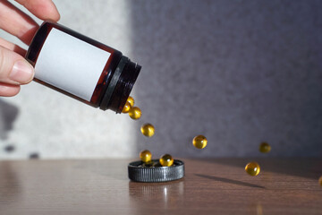 Hand holding pill bottle with yellow capsules spilling on wooden surface in sunlight.
