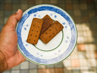 A man's hand carries a plate with several chocolate biscuits on it against a blurry ceramic tile background. Chocolate biscuits with granulated sugar for breakfast.