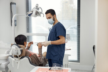Middle aged Caucasian woman sitting in dental chair interacting with young adult Middle Eastern male dentist wearing mask and gloves in modern dental office