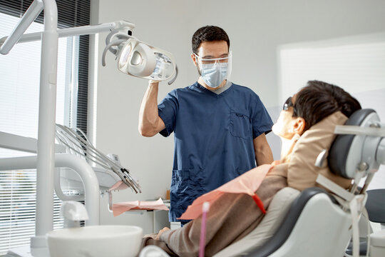 Middle Eastern male dentist preparing dental examination for Caucasian female patient reclining in dental chair, dentist adjusting overhead examination light, patient wearing protective glasses