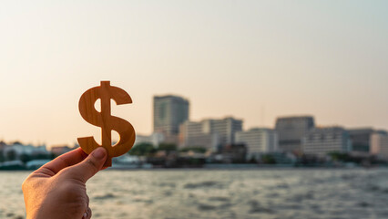 Hand holding wooden dollar symbol with blurred cityscape and river background, representing urban investment, financial growth, economic value, banking, business success, and real estate concept.