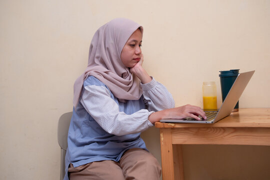 A woman in a hijab works on her laptop with a thoughtful expression, staying focused at her home workspace.