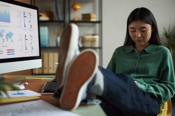 Young adult Asian woman sitting at desk with legs up, looking down at smartphone, working in modern office with computer monitor displaying business analytics charts and graphs
