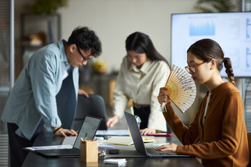 Young Asian businessmen and businesswomen working together in modern office, young Asian woman sitting at desk using laptop and holding paper fan, two young Asian colleagues discussing documents
