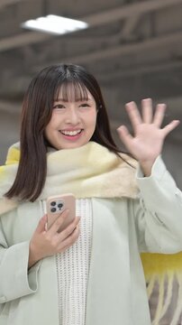 Winter New Year's homecoming season: Close-up of a cute woman wearing a scarf and holding a smartphone on a station platform or bus terminal, waving goodbye in a slow vertical video.