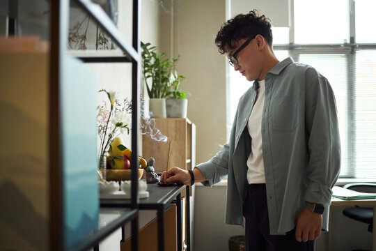 Young adult Asian man standing in modern office using smartphone, wearing glasses and smart watch, working near shelf with decorative items and plants, natural light coming through window