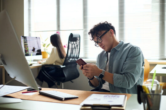 Young Asian man sitting at desk using smartphone while young Asian woman working at computer in modern office workspace, both focused on business tasks and technology