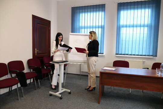 Two women in an office stand by a flipchart, reviewing documents and discussing data during a work presentation.