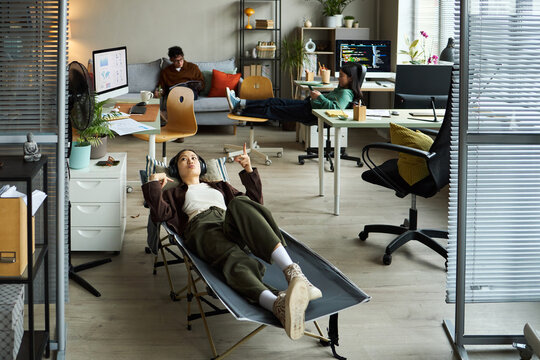 Young Asian businessmen and businesswomen working in modern office, young Asian woman relaxing on lounge chair in foreground while young Asian man and young Asian woman using laptops in background