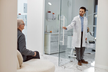 Middle Eastern male dentist opening glass door inviting senior Caucasian man patient into dental office, dental chair and equipment visible in background, both engaging in eye contact