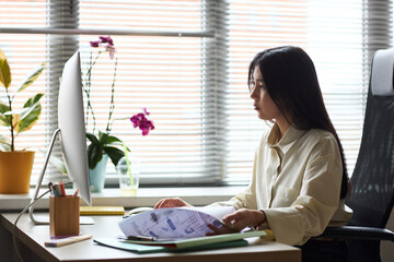 Young adult Asian woman working at desk using desktop computer and reviewing documents in modern office setting, focused on screen while holding paperwork and sitting in ergonomic chair