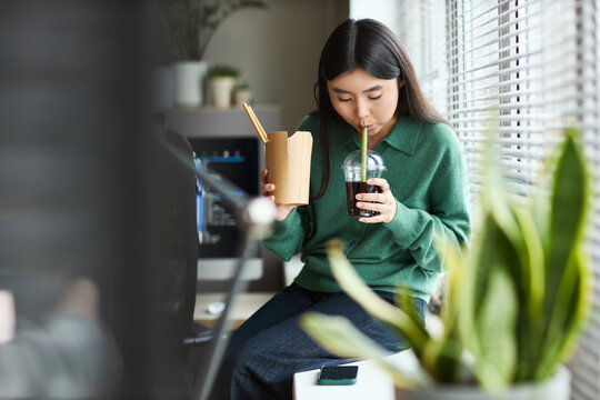 Young adult Asian woman sitting in modern office, eating noodles with chopsticks and drinking iced beverage from plastic cup during lunch break near window with smartphone nearby