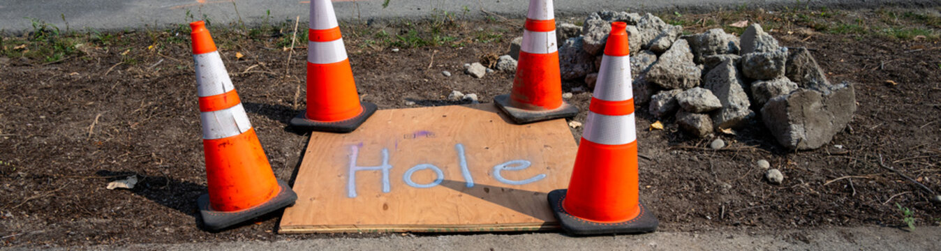 Plywood with the word Hole spay painted on it covering an open hole between the road and sidewalk for underground utilities construction work

