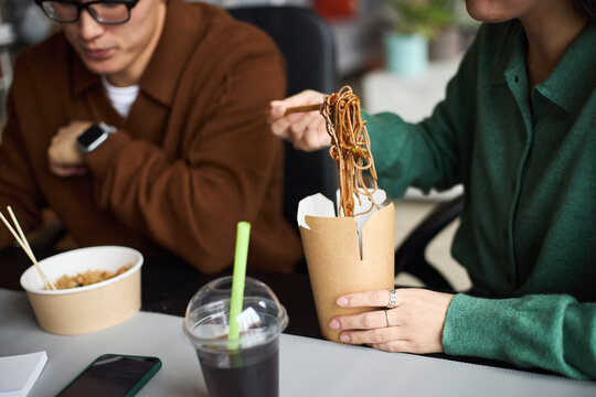 Young Asian man and young Asian woman working in office, eating takeout noodles and rice at desk, using chopsticks, takeaway drink with straw on table, collaborating during lunch break