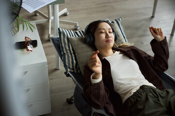 Young adult Asian woman reclining on office lounge chair listening to music with wireless headphones, eyes closed, relaxing during work break, desk and office supplies in background