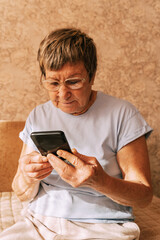 An elderly woman wearing glasses was intently immersed in her smartphone while sitting on a sofa in a sun-drenched. Digital Phone Addiction in Older Women, Screen Time Overload, and Modern Technology