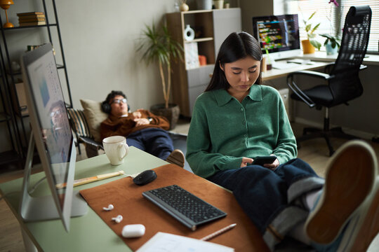 Young Asian woman sitting at desk using smartphone with legs on table, while young Asian man lying on floor in background wearing headphones in modern office workspace setting