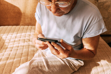 An elderly woman wearing glasses was intently immersed in her smartphone while sitting on a sofa in a sun-drenched. Digital Phone Addiction in Older Women, Screen Time Overload, and Modern Technology